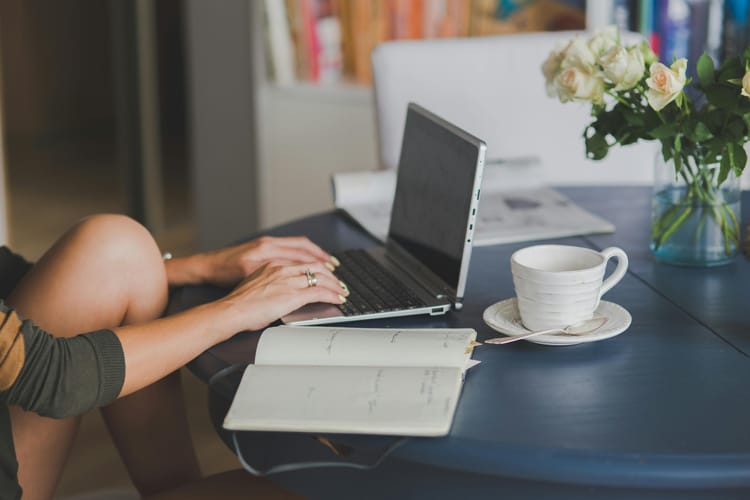 alt="a woman typing on a laptop, sitting at a table with a cup, a notebook and roses"